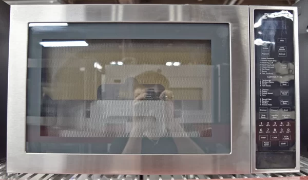 Front view of a brand new Jenn-Air Stainless Steel Series JMC3415ES 25-inch Countertop Convection Microwave Oven with a photographer's reflection on its glass door.